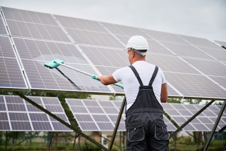 Professional worker cleaning solar PV panel. Man making sure solar batteries in good condition. Back view of worker in overalls and helmet cleaning panel by mop.
