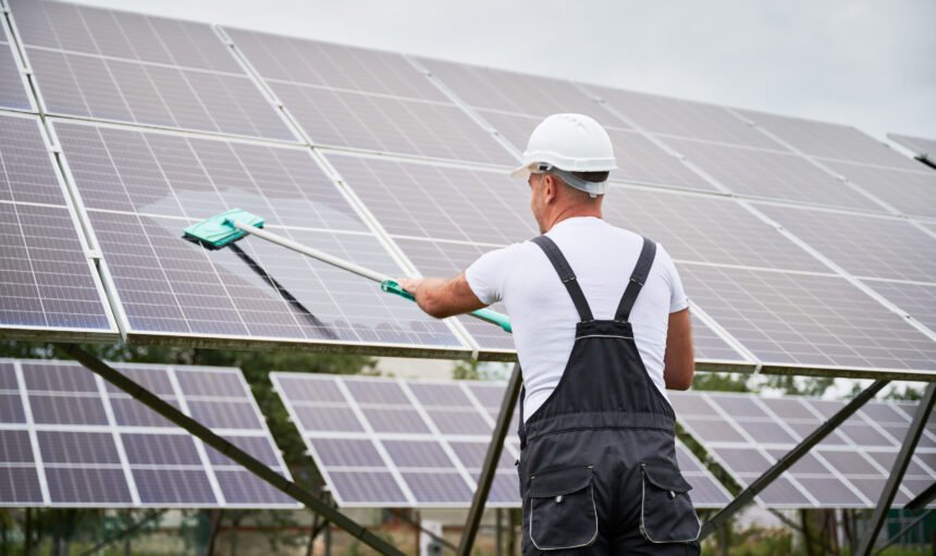 Professional worker cleaning solar PV panel. Man making sure solar batteries in good condition. Back view of worker in overalls and helmet cleaning panel by mop.