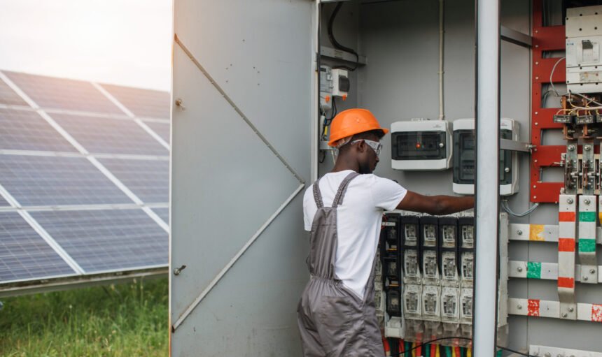 African american man in grey overalls and orange helmet repairing cables in switchgear at solar station. Concept of people, maintenance and alternative energy.